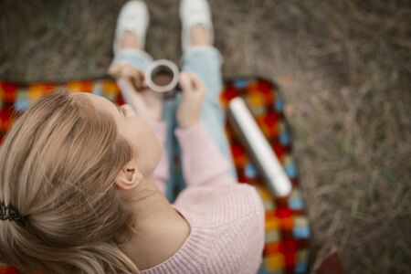 Young Woman Drink Coffee Or Tea In Nature Outdoors, Blonde Lady Holds Cups With Hot Drink Sitting On Picnic Mat Among Grass, Selective Focus On Female Ear, Picnic In Nature Concept, High Angle Viewの写真素材