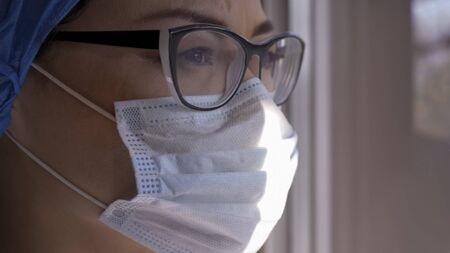 Profile View Of Female Medic Looks At Window In Sunny Day, Portrait Of Sad Young Woman In Protective Mask Forced To Be Isolated During Coronavirus Pandemic, Copy Space At Right Sideの写真素材