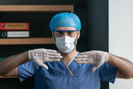 Male Doctor Shows Slogan To Stop Covid Virus Written With Black Marker On His White Gloves, Man In Blue Medical Uniform wants To Stop Covid-19 Or Coronavirus Epidemic, Quarantine Conceptの写真素材