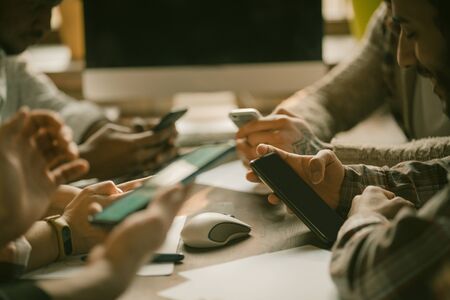 Group Of Young People Using Smart Phones, Close Up Of Human Hands Holding Mobile Phones While Sitting At Office Table Near Window, Side Viewの写真素材