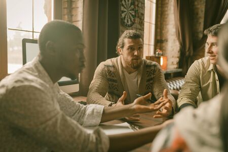 Creative Multi-Ethnic Team Of Freelancers Discussing Startup Project, Colleagues Arguing Each Other Sitting At Table And Gesticulating With Hands In Backlit, Focus On Caucasian Manの写真素材