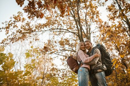 Happy Couple Standing In A Hug Outdoors, Hugging Tourists In Love Stands On Background Of Colorful Autumn Trees And Blue Sky, View From Belowの写真素材
