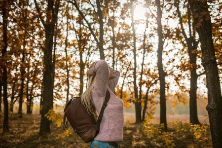 Tourist Woman Traveling In Nature, Young Caucasian Woman With Backpack Arched Her Back With Hands Behind Head And Set Face To The Sun While Standing Against Backdrop Of Autumn Trees On Sunny Dayの写真素材