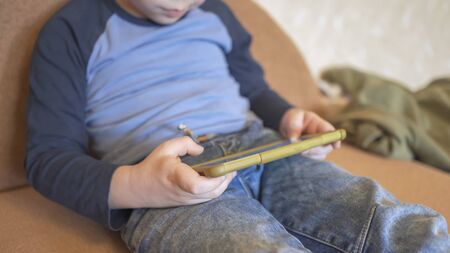 Boy Uses Phone Sitting On The Couch, Caucasian Boy In Casual Wear Spends Time Playing Or Learning...