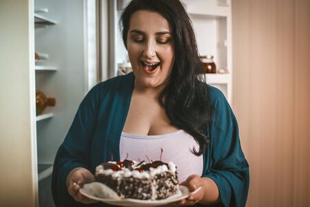 Smiling Overweight Brunette Admires Looking At Cake, Body-Positive Woman Holds Plate Of Sweet...