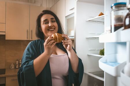 Happy Body Positive Woman Going To Eat Croissant Standing Near An Open Refrigerator In Home Kitchen Interior, Selective Focus On Croissant In Female Handsの写真素材