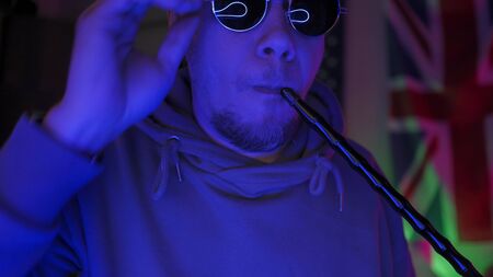 Hipsters Smoking Hookah Having Fun Self-Isolation Time At Home In Neon Backlight, British Man In Sweatshirt Corrects Sunglasses Looking At Camera Against The Background Of Great Britain Flagの写真素材