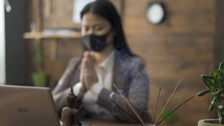 Prayer Of Asian Business Woman Sitting In Mask On Her Face At Office Table In Front Of Laptop, She Clasped Hands Together Reading Prayer, Business During The COVID 19 Pandemicの写真素材