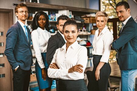 Multi-Ethnic Group Of Office Worker Posing Together, Business Colleagues In Formalwear Smiles Standing In Office, Selective Focus On Woman With Crossed Hands In Foreground, Toned Imageの写真素材