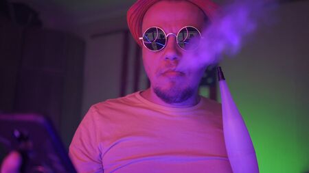 Stylish Man In Sunglasses Uses A Mobile Phone Smoking A Hookah, Guy Reading News On Self-Isolation Is Sitting In Home Interior With A Neon Glow, Survive The Quarantine Conceptの写真素材