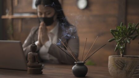 Woman in a face mask is meditating at Office workplace. Focus is on incense sticks and a Buddha figurine standing on a wooden table in the foreground.の写真素材