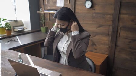 Asian woman corrects a protective mask before working with a computer. Oriental Woman Works Alone On Laptop In Office Interior. Concept of quarantine and social distance.の写真素材