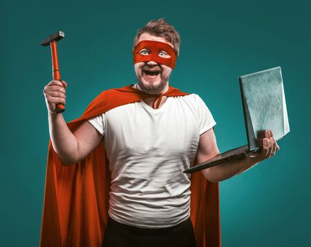 Smiling Super hero man holds hammer and laptop ready to repair the computer. Man In Red Super Hero Uniform posing on biscay green background. Computer repair concept.の写真素材