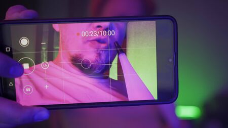 Man Makes Self Video with Mobile Phone Smoking Hookah In Neon Light. Focus on the screen of a...