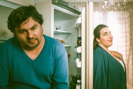 Thoughtful body-positive couple stands near the open fridge in home kitchen, a man and a woman in blue casual clothes dream together.の写真素材