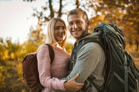 Happy couple of backpackers hugging while looking at the camera, a Caucasian couple in love toothy laughs while standing against the backdrop of colorful autumn nature.の写真素材