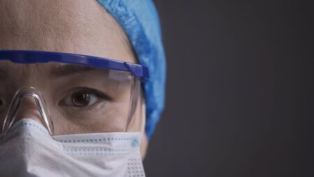 Tired doctor looks at camera. Close up shot of medic eye. Serious Female nurse wearing protective mask, cap and protection glasses. Copy space at right side on grey background.の写真素材