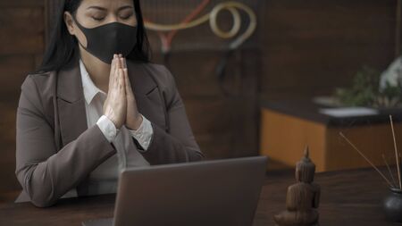 Asian woman prays sitting at workplace in oriental style with incense sticks and buddha statuette on a wooden wall background. Meditation concept.の写真素材