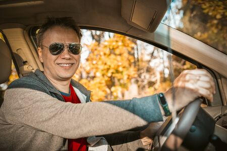 Driver stopped car in autumn forest. Handsome man in sunglasses smiles at the camera sitting in the car against the background of trees with yellow foliage outside the window.の写真素材
