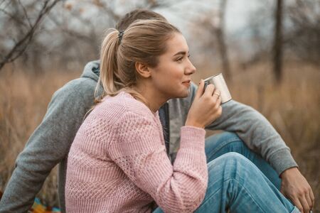 Picnic in nature. Young woman and man rests drinking hot coffee or tea while sitting on blanket at autumn grass outdoors. Profile view. Lifestyles concept.の写真素材
