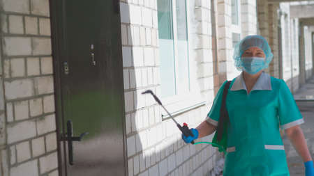 Worker holds a device disinfector looking at camera, standing against brick wall at entrance to building on sunny day. Cleaning concept. City hospital. May, 2020, Brovary, Ukraine.のeditorial素材