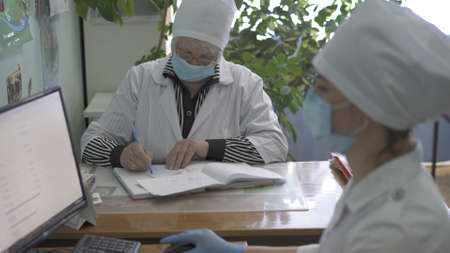 Work of doctors team in ophthalmologist office. Selective focus on mature doctor writing medical forms, young nurse works with a computer in foreground. City Hospital. May, 2020, Brovary, Ukraine.のeditorial素材