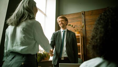 Business handshake of young businessman and gray-haired businesswoman looking at each other standing at office desk after team brainstorming in office. Shot from below.の写真素材