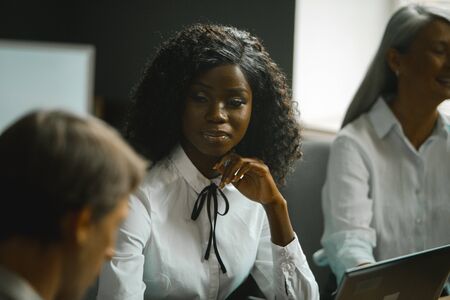 Diverse team brainstorming in office. Highlighted focus on African-American young woman sitting at table along with grey-haired mature woman and Caucasian man.の写真素材