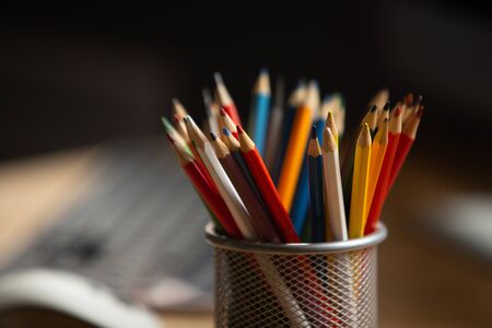 Office workplace. Focus on multi-color pencils in foreground at wooden table with laptop computer on it. Copy space for text at left side. Close up shot.の写真素材