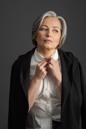 Charming gray-haired woman thoughtfully looks away adjusting collar of white shirt, throwing black jacket over her shoulders. Isolated on gray background. Lonely concept.の写真素材