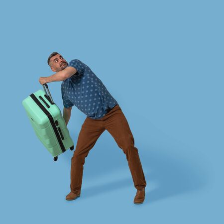 Man holds plastic suitcase posing in studio on blue background. Caucasian man going to throw his green suitcase with luggage upstairs. Travel advertising template with textspace.の写真素材