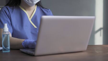 Medic working laptop sitting at table with computer and antiseptic spray on it in blurred foreground. Highlighted focus on female torso in blue uniform. Close up shot. Copy space at right side.の写真素材