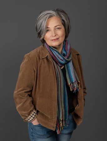 Pretty gray-haired woman in casual. Lovely mature lady wearing brown jacket and colorfull scarf standing hands in pockets on on gray background. Female studio portrait.の写真素材