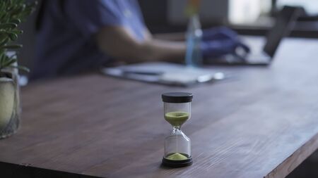 Hourglass on desk of doctor working with computer. Close up shot. Selective focus on hourglass with...