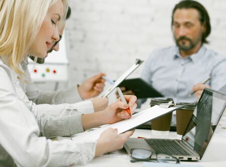 Business team brainstorming in office. Colleagues works sitting at office table with laptop and coffee cups on it. Selective focus on business woman writing by red marker on paper sheet in foreground.の写真素材
