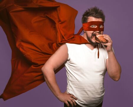 Hungry superhero man eats big hamburger with meat. Man in red flying cloak eats looking at camera on grape purple background. Fast food snack concept.の写真素材