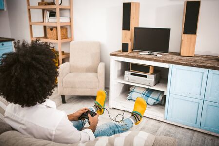 Stylish guy plays computer games sitting on couch. Young unrecognizable man with afro hairstyle in yellow slippers relaxing in home interior. Home alone concept. Rear view. Toned image.の写真素材