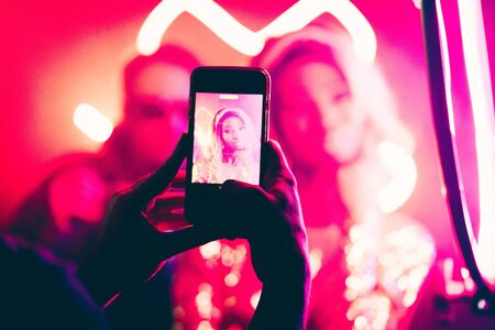 Young woman is photographed in a night club. Focus on male hands making photo by smartphone in...