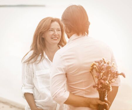 Dating of happy mature couple on seashore. Man about to give bouquet of flowers standing with his...