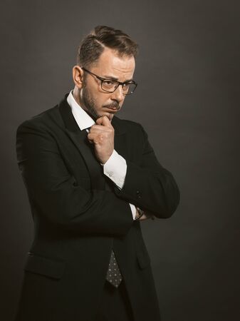 Doubting businessman crossed his arms touching his chin. Thoughtful intelligent man in glasses stands hesitantly looking to the side. Doubt concept. Toned image.の写真素材