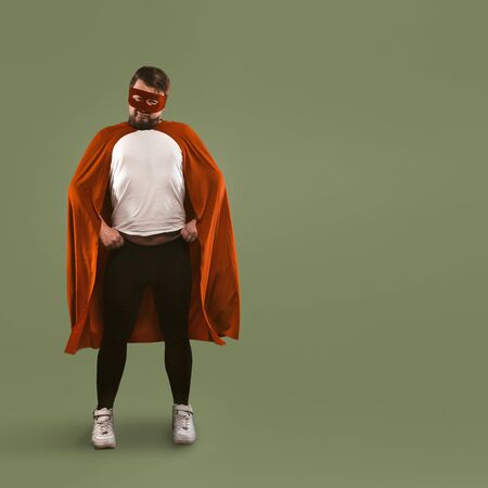 Superhero man stands akimbo at full height. Young Caucasian man in a red superhero costume looks at camera. Isolated on green background. Template with place for text.の写真素材