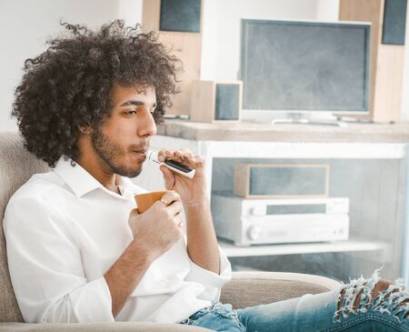 Young man drinks coffee smoking of electronic cigarette sitting at home interior against the background of TV and sound system. Rest concept. Tinted image.の写真素材
