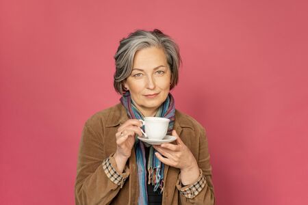 Charming Caucasian woman drinks coffee holding white cup. Pretty mature lady smiles slightly isolated on pink background. Beauty concept. Coffee time concept.の写真素材