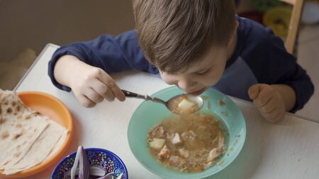 Cute preschool child learns to eat by himself. Hungry boy holds big spoon eating soup sitting alone at table. High angle view.の写真素材