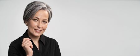 Pretty mature woman smiles with hand near face. Charming gray-haired lady in black blouse on white...