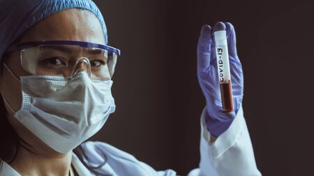 Female lab worker holds blood test tube looking at camera. Asian woman in protective mask and goggles on gray background. Vaccine concept. Close up shot. Tinted image.の写真素材