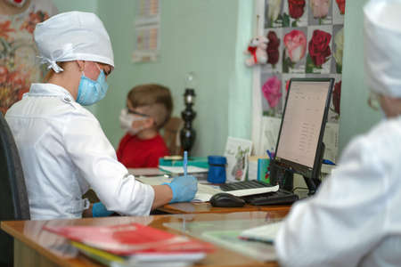 Doctor makes notes on card of little patient after examination, recording appointment and results. Masked woman and boy due to virus outbreak and pandemic. City Hospital. May, 2020, Brovary, Ukraine.のeditorial素材