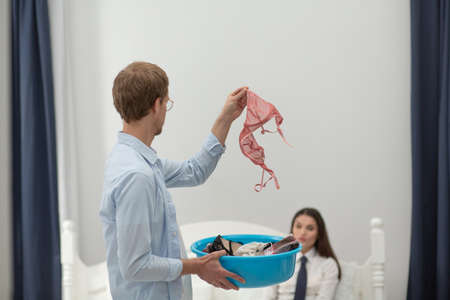 Man holding up his partners lingerie in his hand. Guy wearing home clothes holding up his girlfriends pink bralette and basin full of lingerie indoors in bedroom.の写真素材