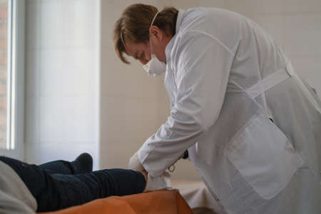 Doctor surgeon examining his patient leg. Medic inspects of patient lying on couch examining his leg. City Hospital. May, 2020, Brovary, Ukraine.のeditorial素材