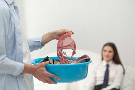 Man holding womans lingerie in his hand. Close up shot of mans hand picking up pink lace bralette out of laundry basin. His girfriend businesswoman blurred in background.の写真素材